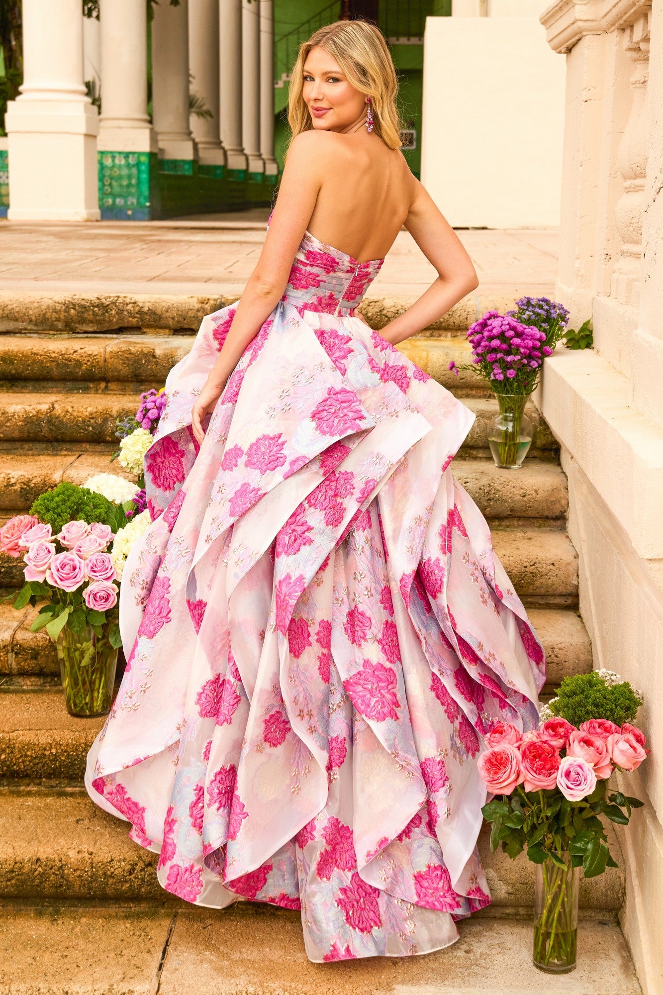 A woman wearing the Ava Presley 42293 Strapless Print Prom Ball Gown stands on outdoor steps with pink and purple bouquets, a classical building in the background.
